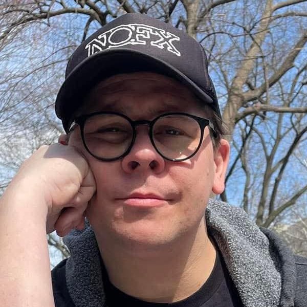 Zach Lucas in a black cap and glasses, resting his chin on his hand at an outdoor chess table